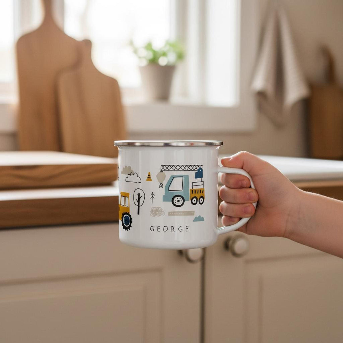 A hand holds up a white enamel mug printed with construction vehicles and the name George in front of a kitchen background with cutting boards.