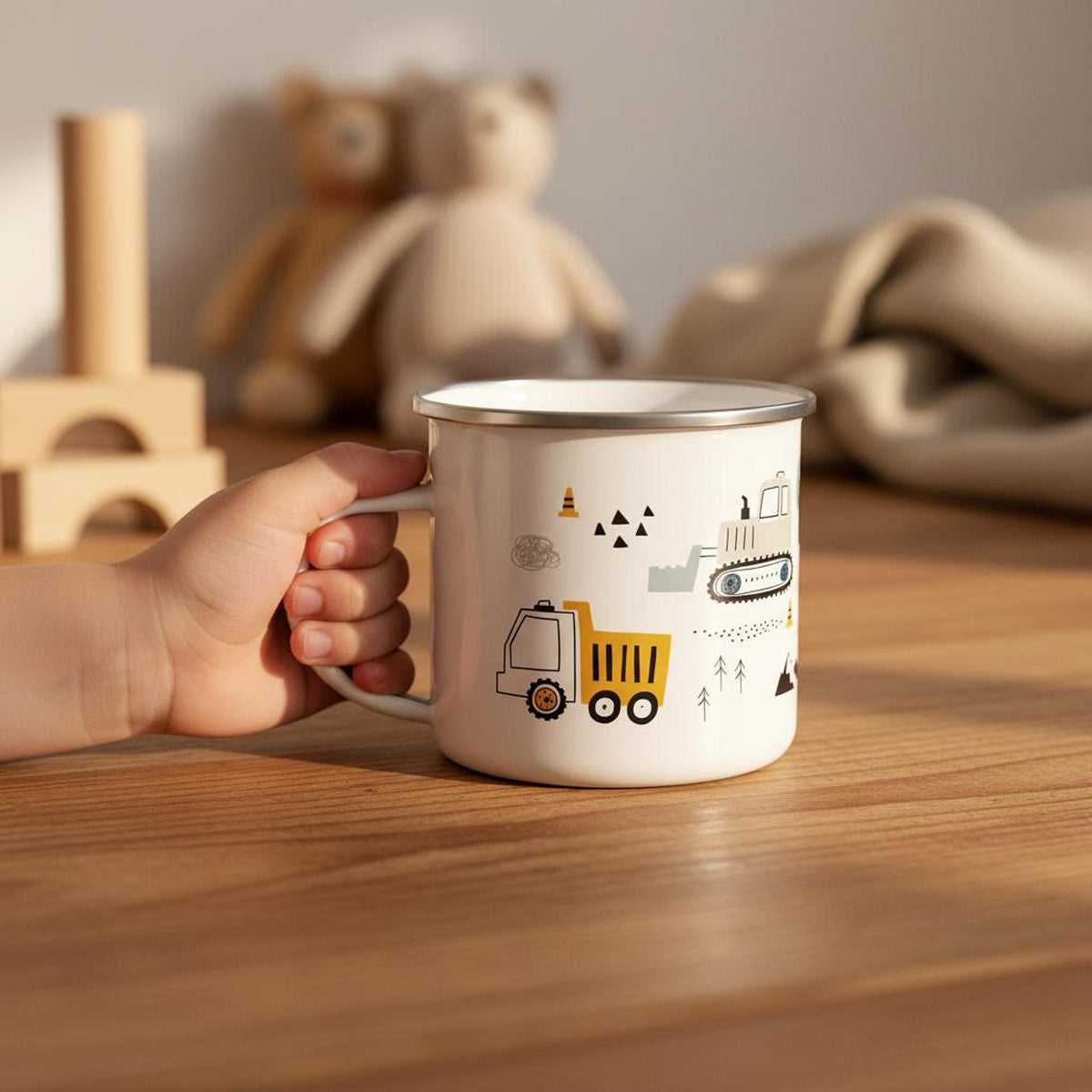 A child's hand holds a white enamel mug with a construction design, resting on a wooden tabletop with wooden toys and a textile background.