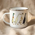 A white enamel mug featuring an illustration of five colorful Indian runner ducks walking in a row, sitting on a textured neutral fabric background.