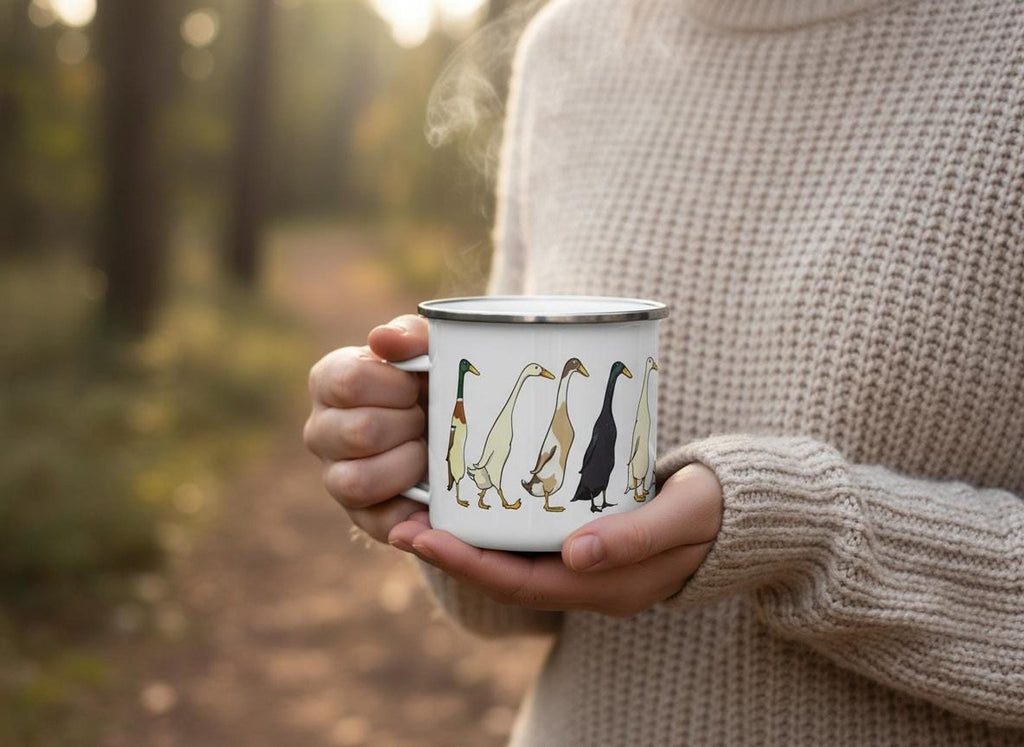 Close-up of a person in a knit sweater holding a steaming white enamel mug printed with runner ducks, with a forest background.