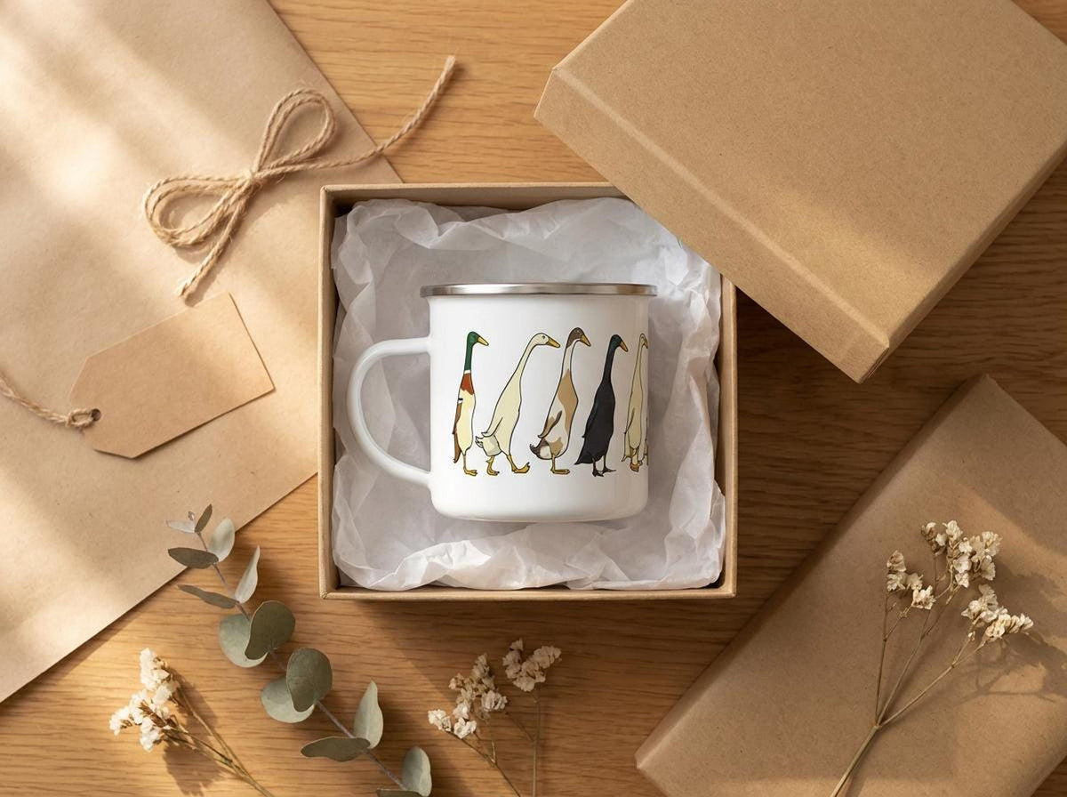 An enamel mug with an illustration of runner ducks sits open inside a cardboard gift box, surrounded by tissue paper, gift wrap, and eucalyptus leaves on a wooden table.