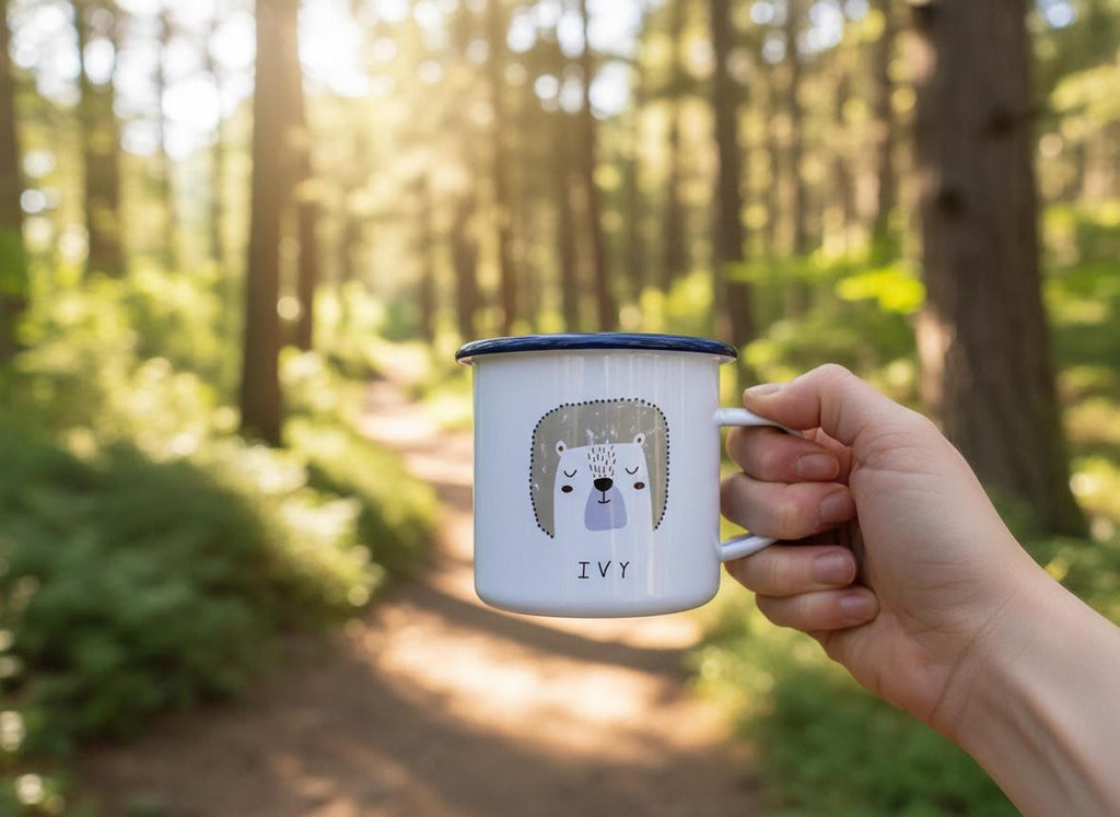A hand holds a white enamel mug with a bear graphic and the name Ivy in a sunlit forest.