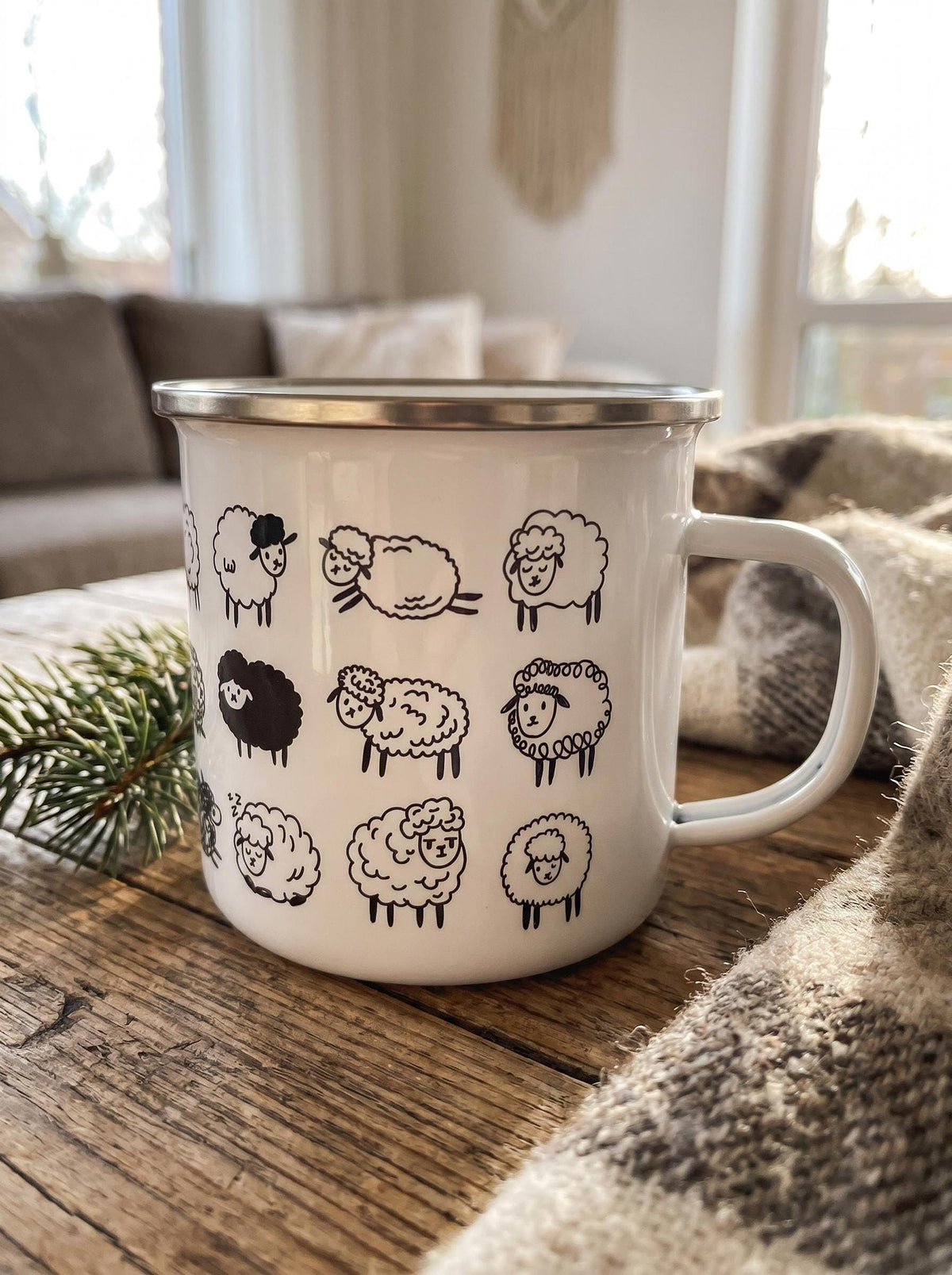 A white enamel mug with black sheep designs sits on a rustic wooden table alongside a pine branch and a patterned blanket.
