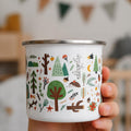 A hand holds a white enamel mug decorated with a colorful woodland pattern of trees, leaves, animals, and mushrooms.