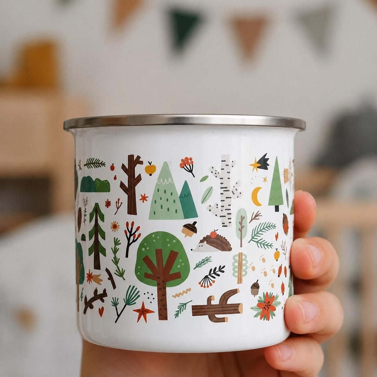 A hand holds a white enamel mug decorated with a colorful woodland pattern of trees, leaves, animals, and mushrooms.