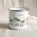 A white enamel camp mug with a silver rim displays a playful illustration of camper vans on a winding road with mountains, trees, and the text TIME TO EXPLORE, resting on a textured linen surface.