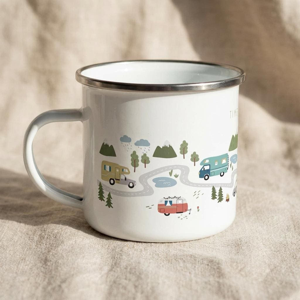 The white enamel mug is turned slightly to show the handle and a continuation of the road trip illustration with colorful vans, lakes, and pine trees against a beige fabric background.