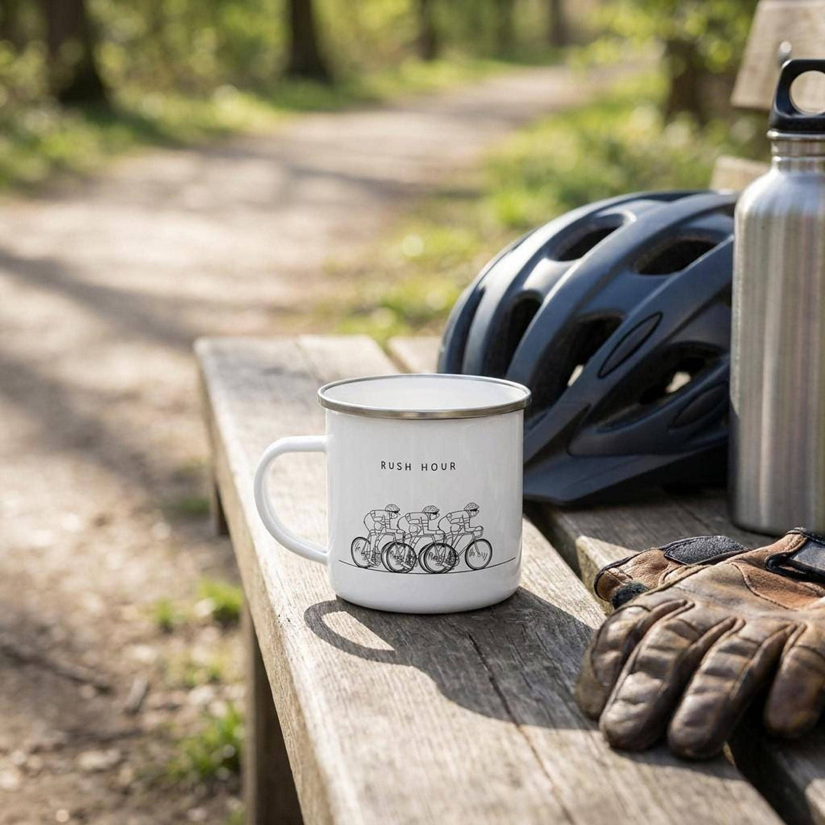 A white enamel mug with the RUSH HOUR cycling design rests on a wooden bench outdoors next to cycling gloves, a helmet, and a water bottle.