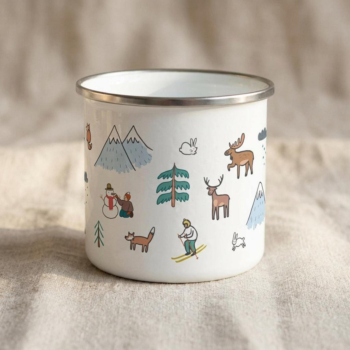 The white enamel mug is rotated to show a different section of the winter motif, featuring moose, foxes, rabbits, trees, and snow-capped peaks against a neutral cloth background.
