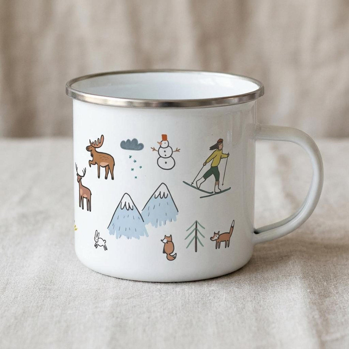 A third angle of the winter-themed enamel mug displays a skier, blue mountains, deer, and a snowman on its white surface.