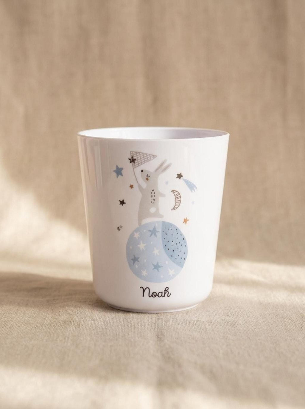 A white polymer cup featuring a blue moon with stars and a grey bunny holding a net, printed with the name Noah, standing on a textured beige surface.