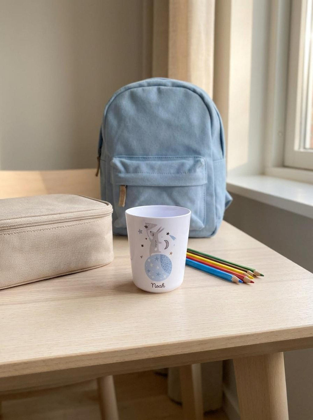 A wooden desk holding the white bunny cup, a light-colored pencil case, colored pencils, and a blue backpack against a bright window backdrop.