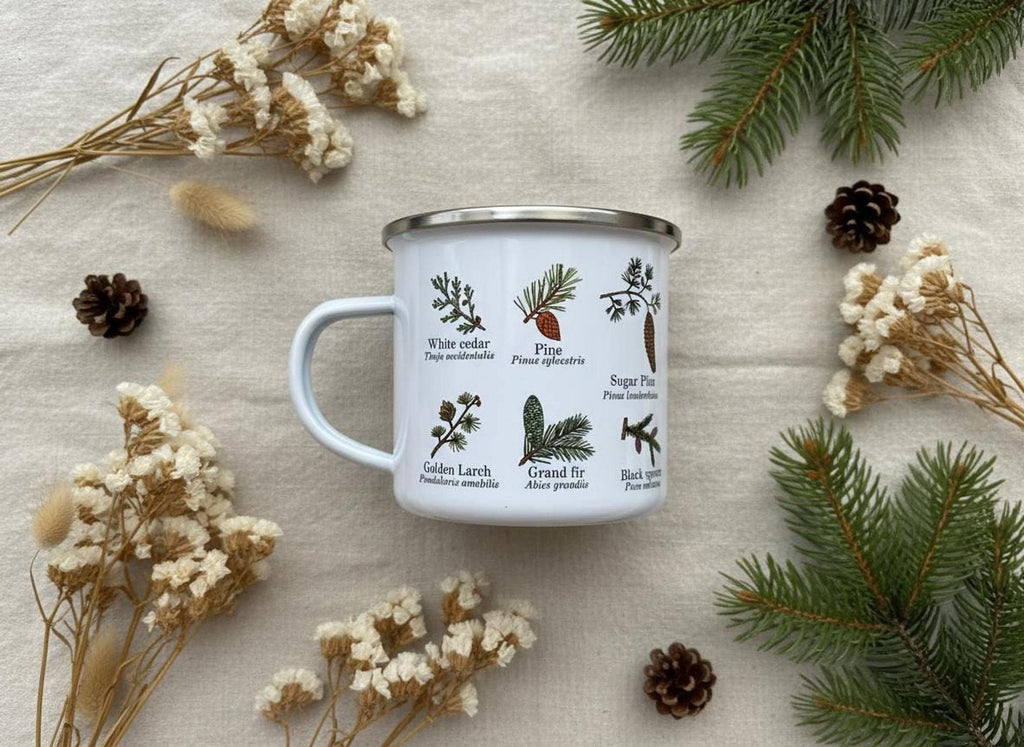 The white enamel mug with conifer tree drawings is surrounded by dried white flowers, pinecones, and evergreen sprigs on a linen surface.