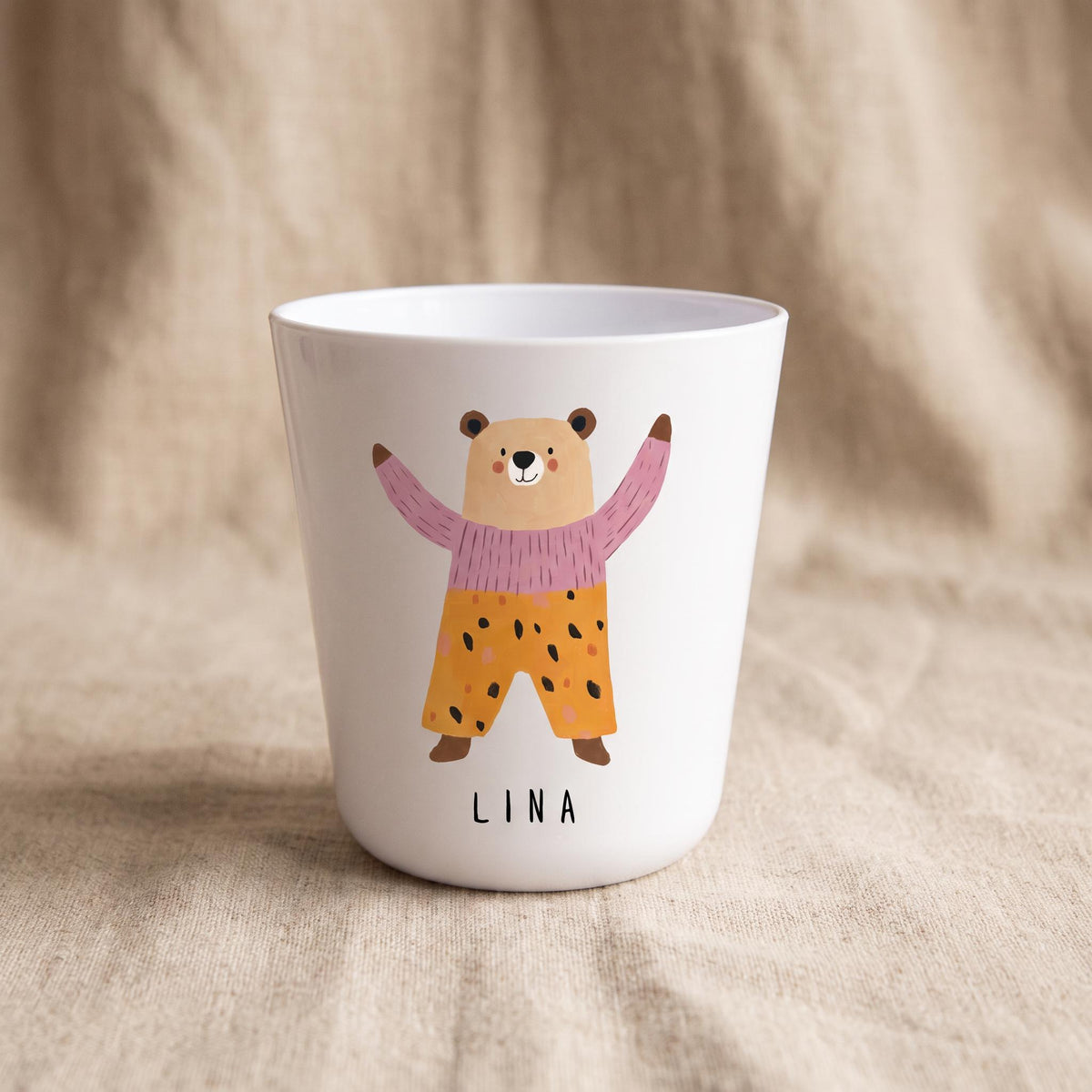 A white polymer cup decorated with a cartoon bear in a pink sweater and yellow patterned pants, with the name Lina printed underneath.