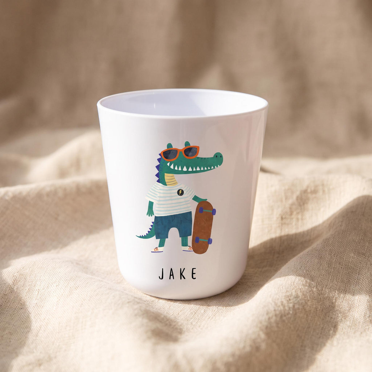 A white polymer cup featuring a crocodile wearing sunglasses and a striped shirt while holding a skateboard, with the name Jake printed underneath.