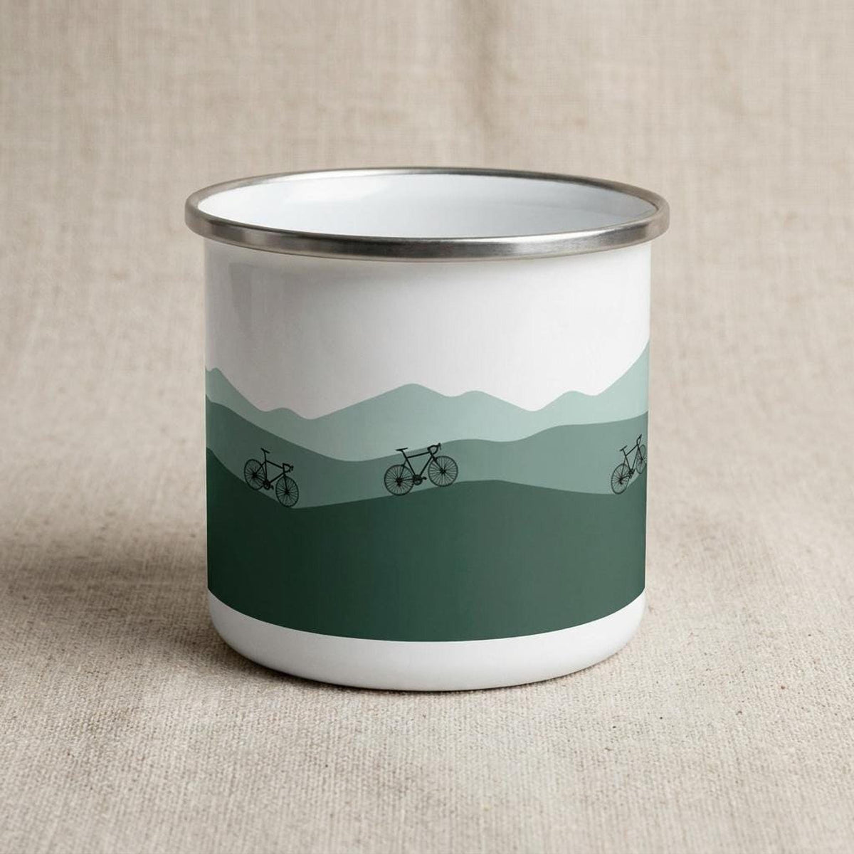 A white enamel mug with a silver rim featuring a green mountain landscape graphic with three bicycles.