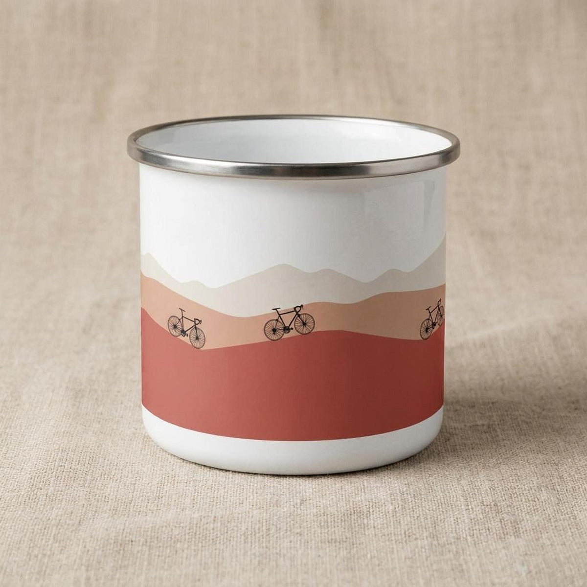 A white enamel mug with a silver rim featuring a red and beige mountain landscape graphic with three bicycles.