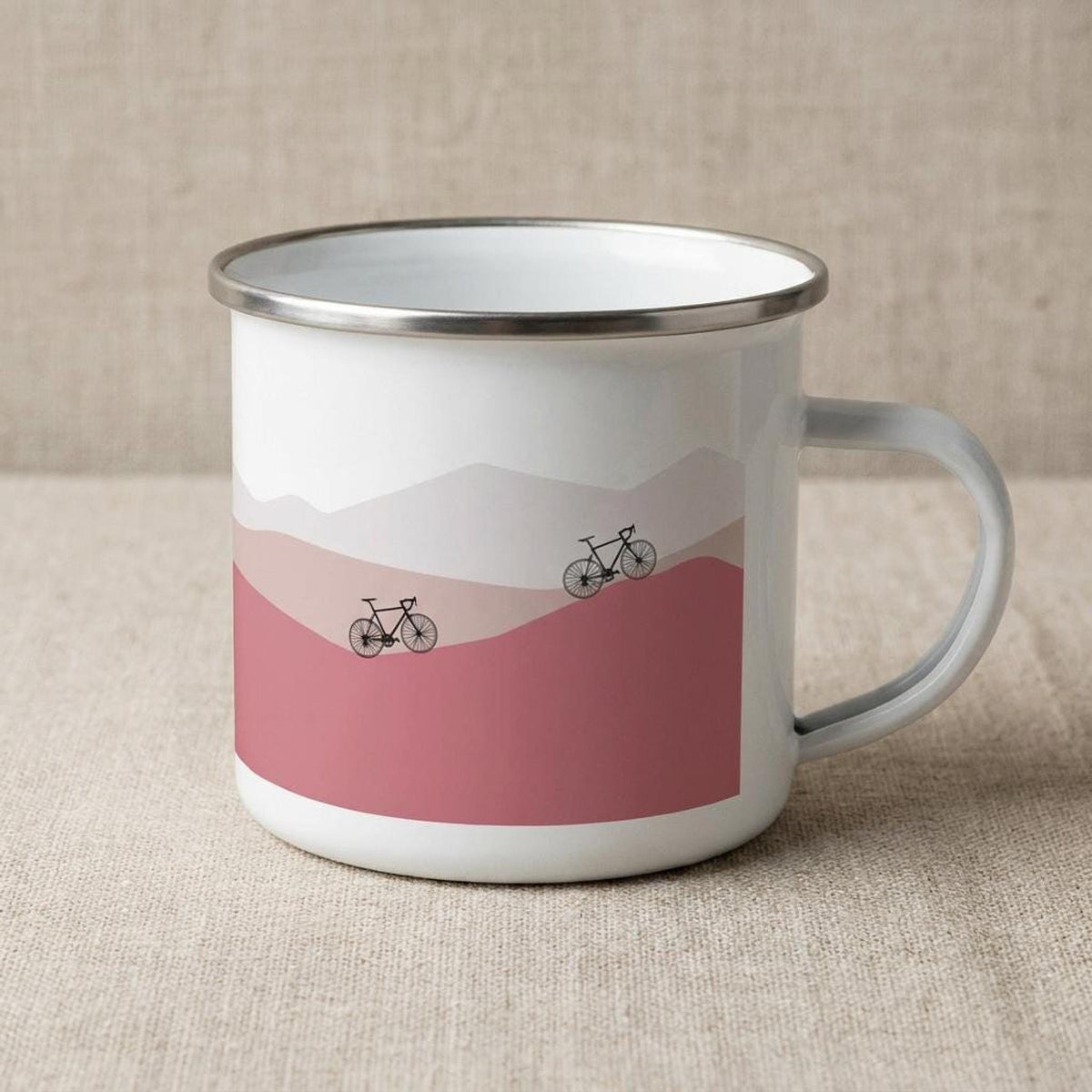 A white enamel mug with a silver rim featuring a pink mountain landscape and two bicycles.