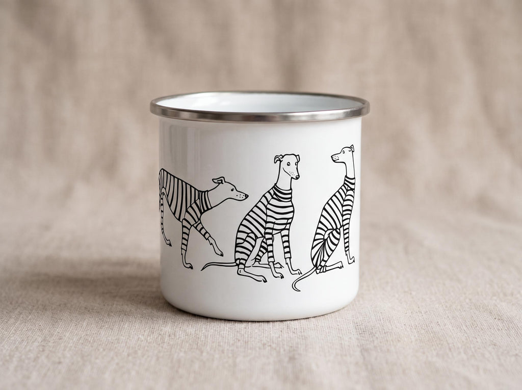 A white enamel mug with a silver-coloured rim featuring a black line art illustration of three greyhound-type dogs wearing striped jumpers. The dogs are depicted in various poses, including walking and sitting, against the white surface.