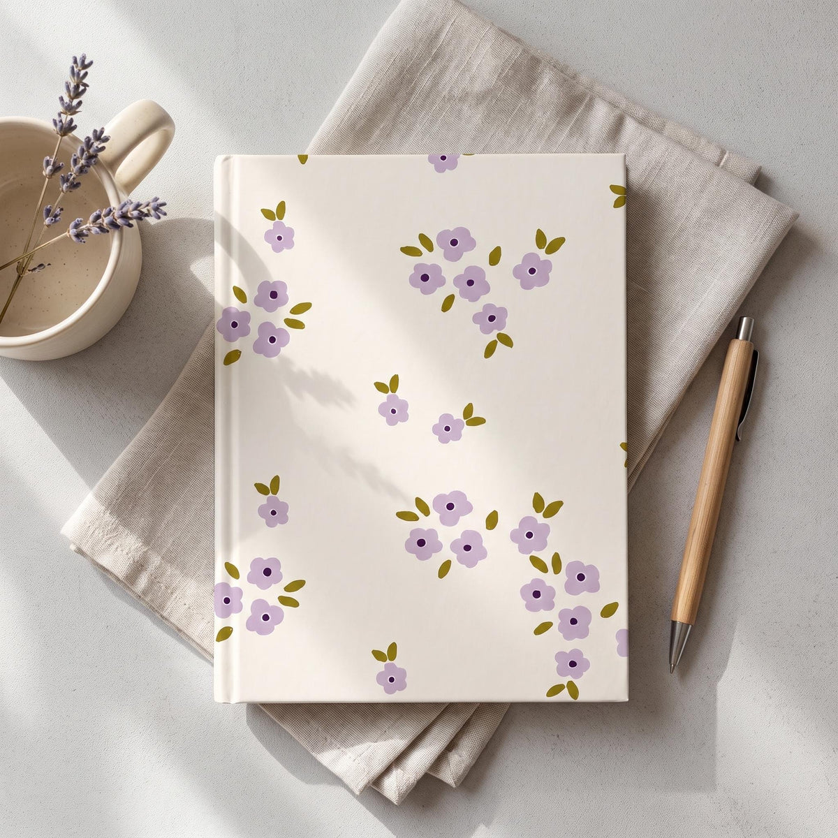 A hardback journal with a cream-coloured cover featuring a repeating pattern of small, light purple five-petalled flowers with dark purple centres and olive green leaves. The journal is positioned next to a wooden pen.