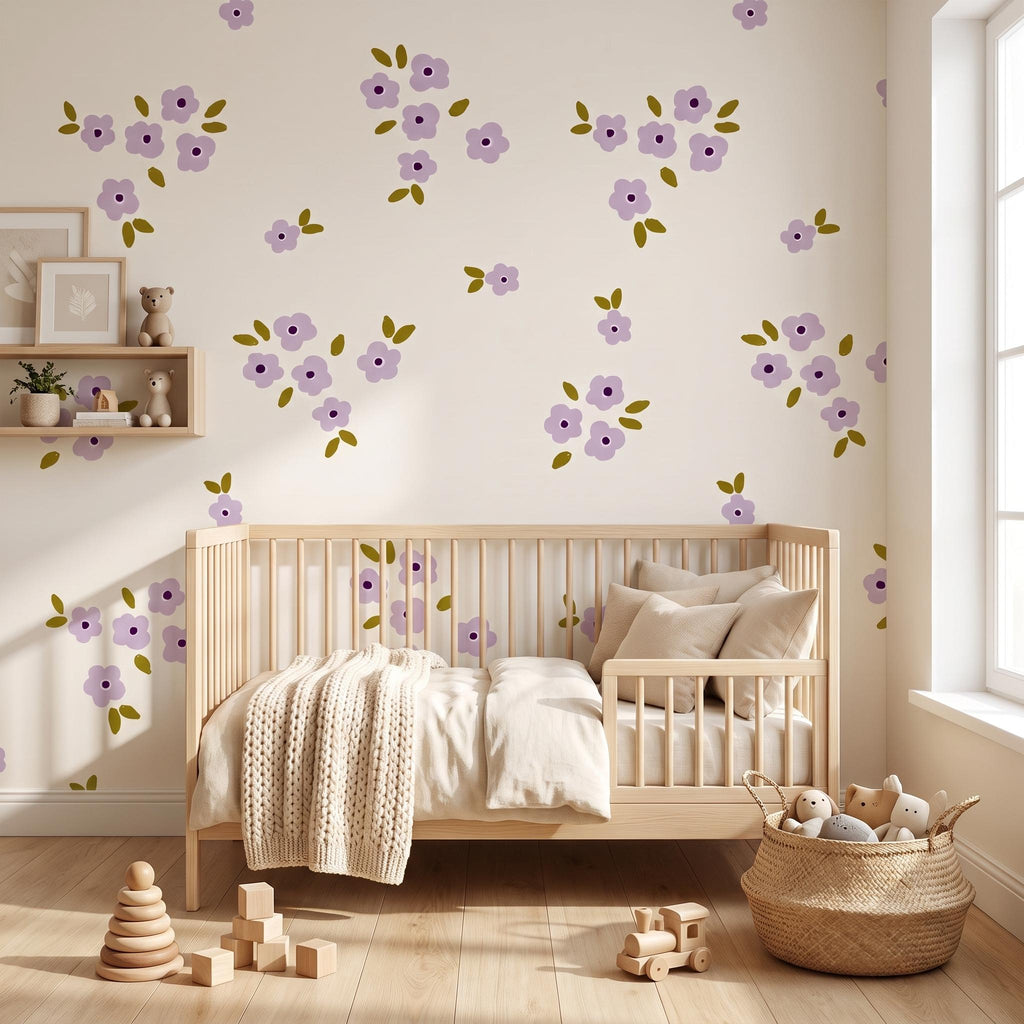 A light wood cot with a cream-coloured blanket and pillows sits against a wall decorated with purple flower decals featuring olive green leaves. A wooden shelf holds small toys, while a woven basket contains plush items on the floor.
