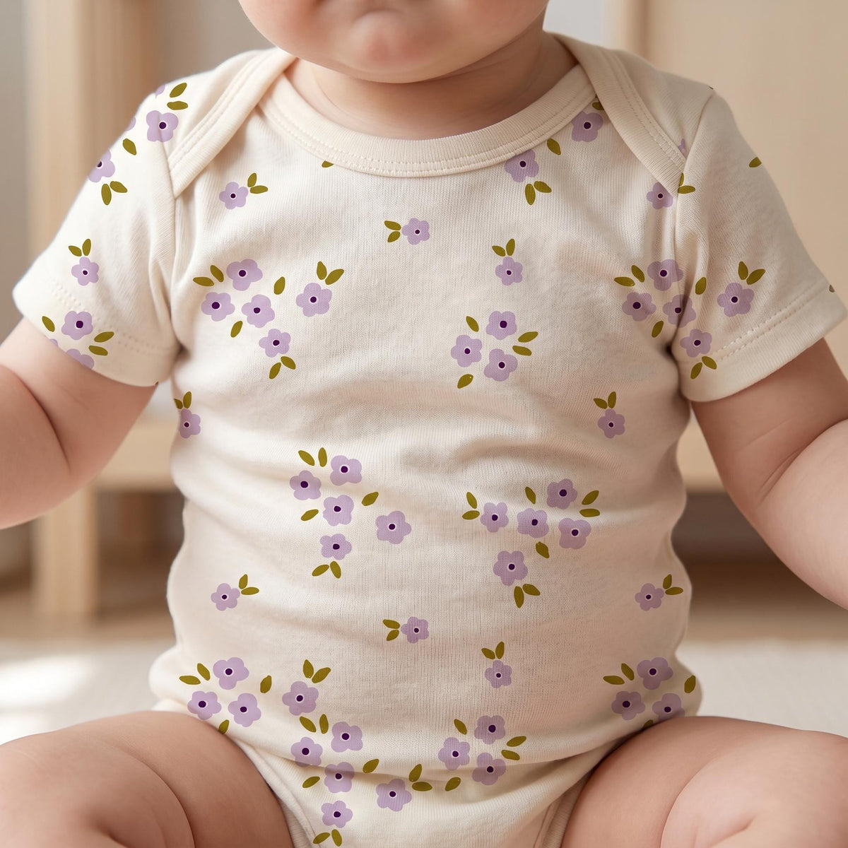 A short-sleeved baby bodysuit in a cream colour featuring a repeating pattern of small, light purple flowers with dark purple centres and tiny olive green leaves scattered across the fabric, including the sleeves and the round neckline.
