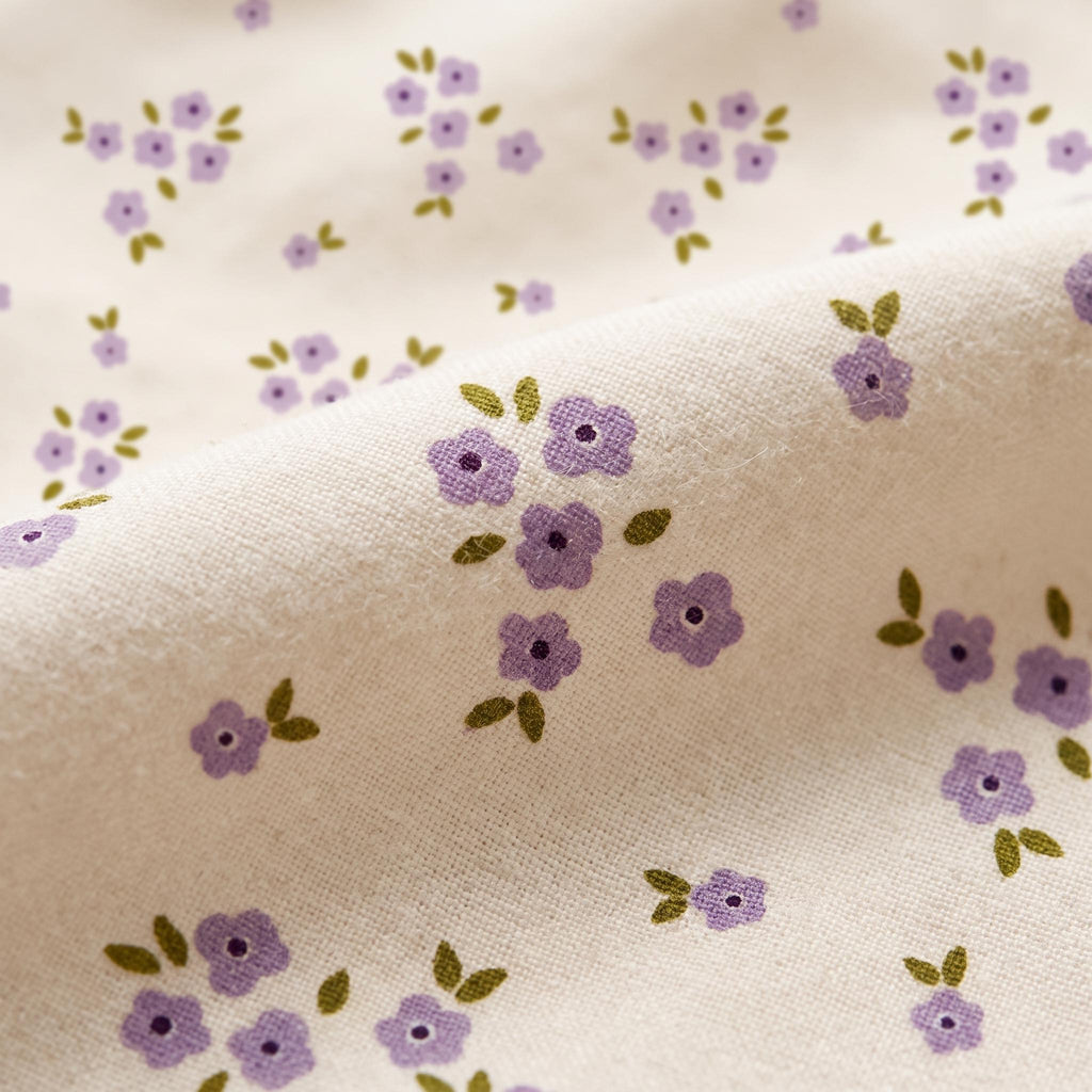A light beige fabric featuring a repeating pattern of small, five-petalled purple flowers with dark purple centres and tiny olive green leaves. The textile has a visible woven texture with soft folds creating depth across the surface.