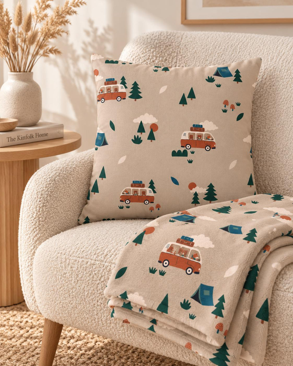 A square throw pillow and a matching folded blanket featuring a repeating pattern of orange camper vans, green pine trees, blue tents, and small red mushrooms on a light beige background, showcasing a camping-themed design.