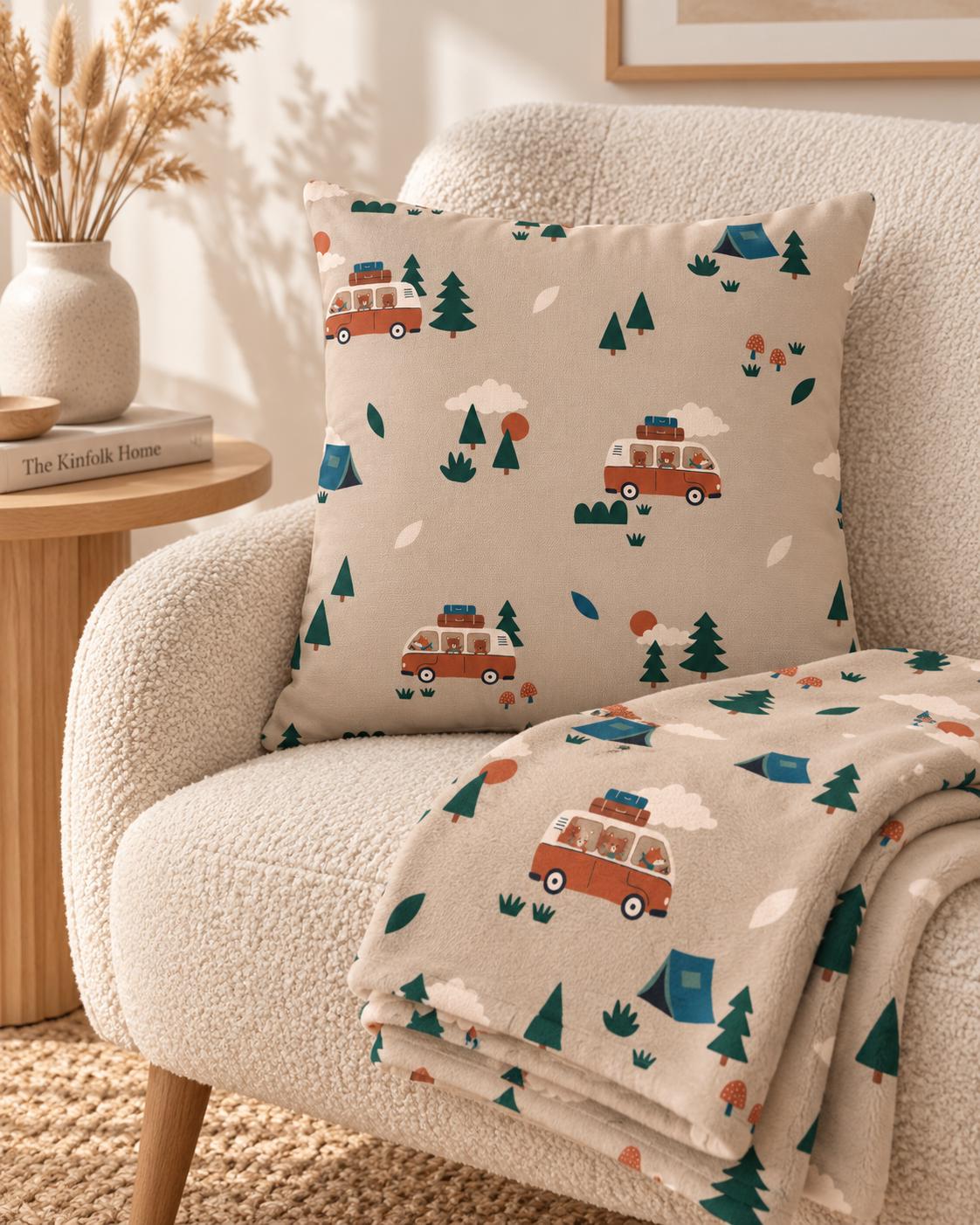 A square throw pillow and a matching folded blanket featuring a repeating pattern of orange camper vans, green pine trees, blue tents, and small red mushrooms on a light beige background, showcasing a camping-themed design.