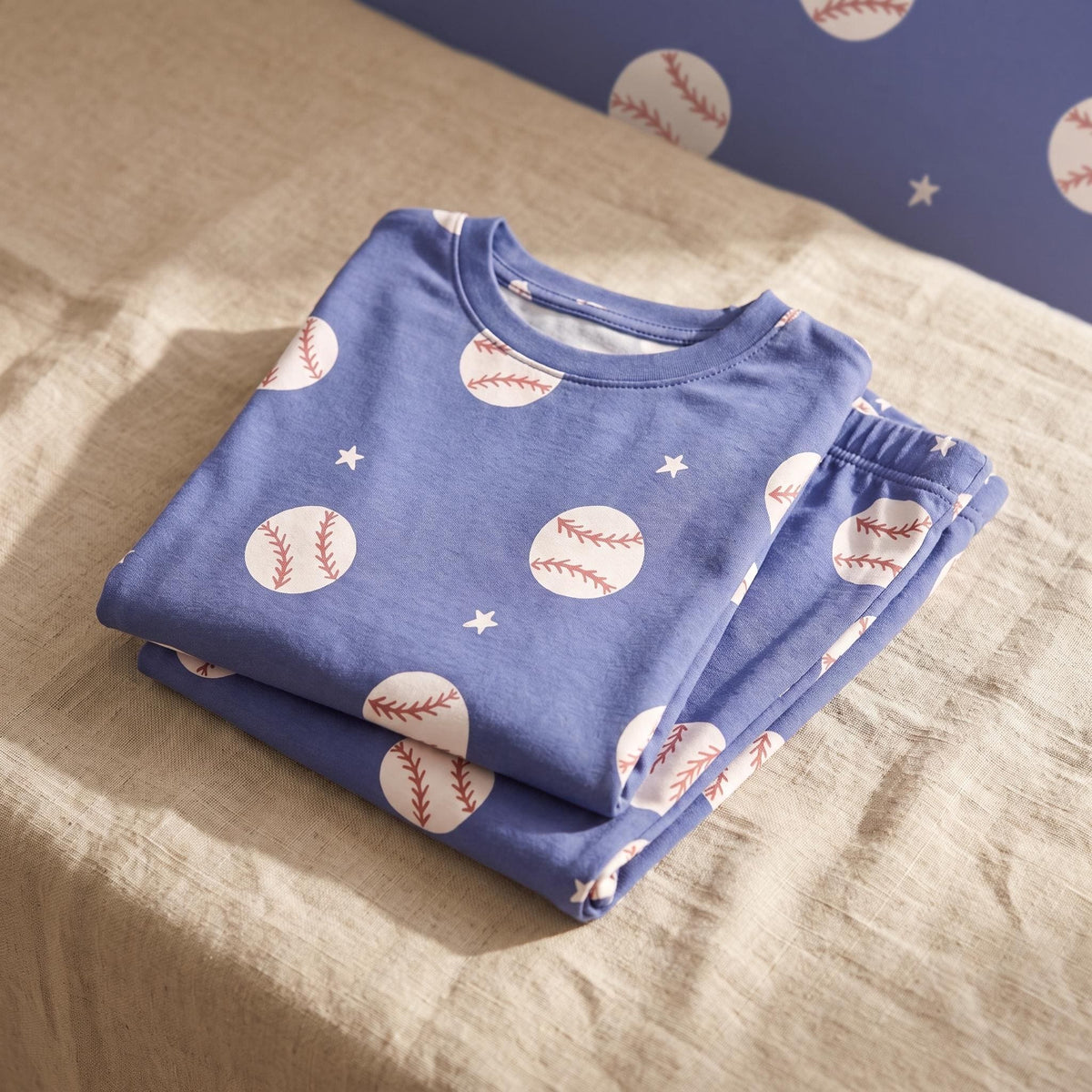 A folded blue long-sleeved shirt and matching trousers set featuring a repeating pattern of white baseballs with red stitching and small white stars. The fabric has a crew neckline and an elastic waistband on the folded trousers.