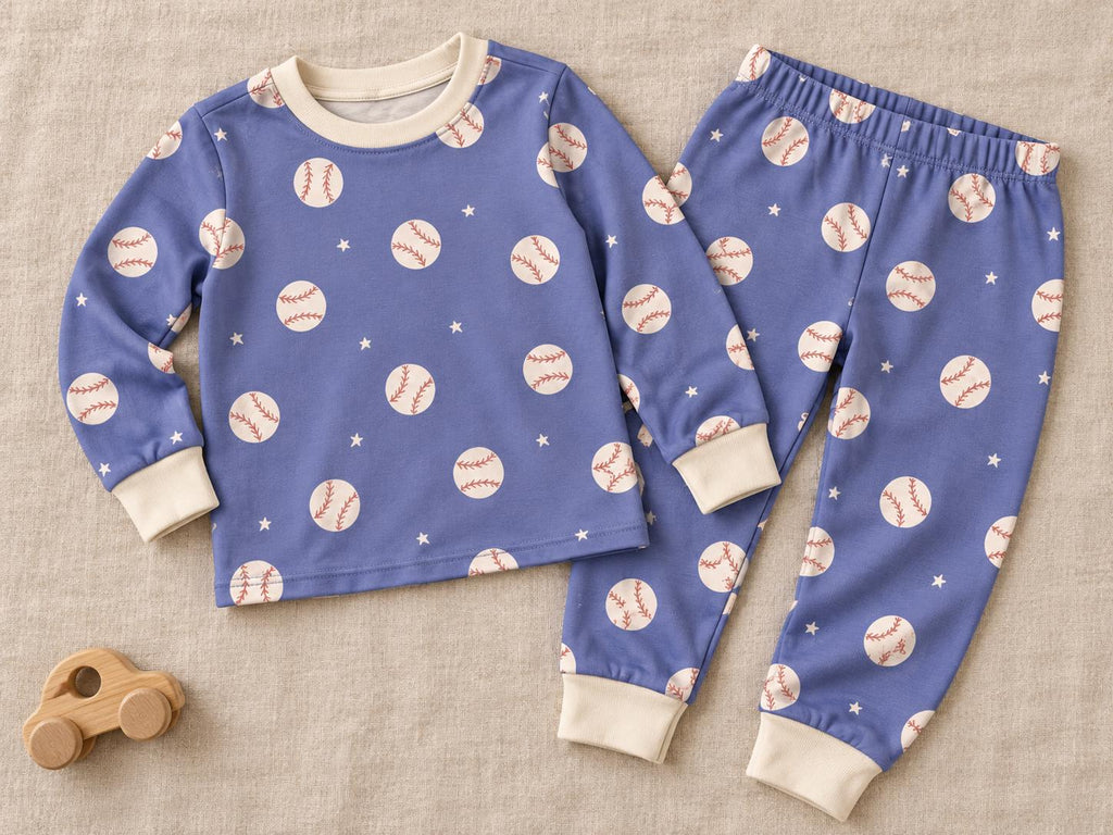A long-sleeve shirt and trousers set in periwinkle blue featuring a repeating pattern of white baseballs with red stitching and small white stars. The garment has a cream-coloured crew neckline and matching cream-coloured ribbed cuffs.