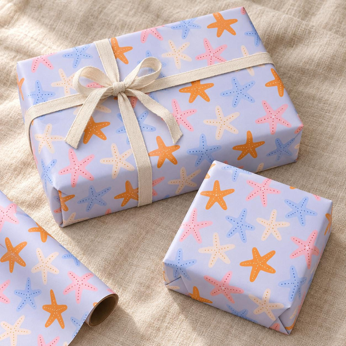 A rectangular gift box and a smaller square gift box wrapped in light purple paper featuring a repeating pattern of orange, pink, and blue starfish. The larger box is tied with a cream-coloured fabric ribbon in a bow.