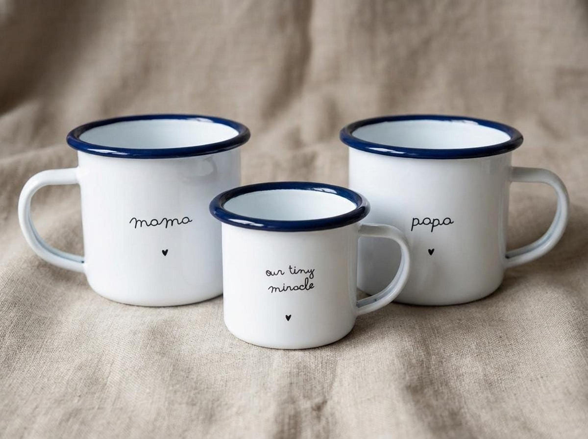 Three white enamel mugs with blue rims displaying mama, our tiny miracle, and papa with small hearts on a burlap background.