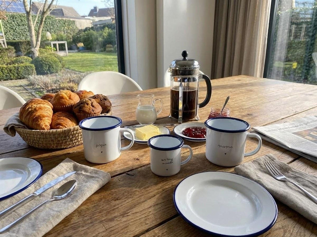 A breakfast table spread with croissants in a basket, french press, plates, cutlery, and three matching enamel mugs reading papa, our tiny miracle, and mama.