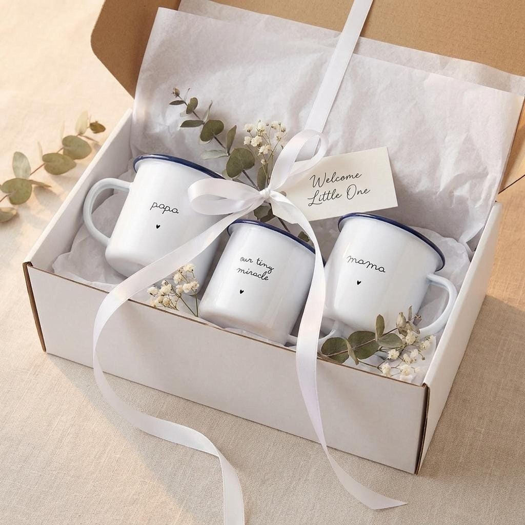 An open white gift box containing three white enamel mugs with blue rims, tissue paper, eucalyptus sprigs, and a Welcome Little One tag tied with a white ribbon.