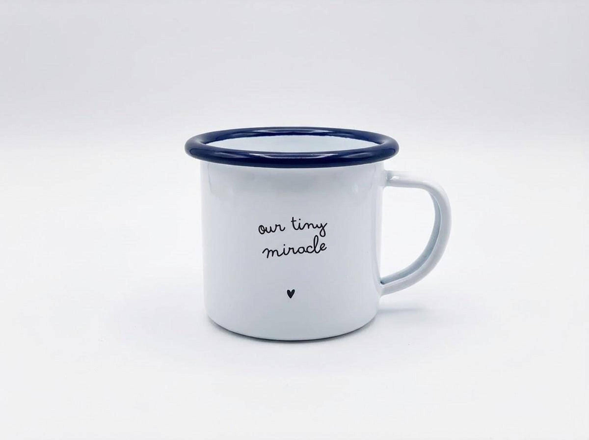 A small white enamel mug with a blue rim featuring the text our tiny miracle and a small heart against a plain white background.