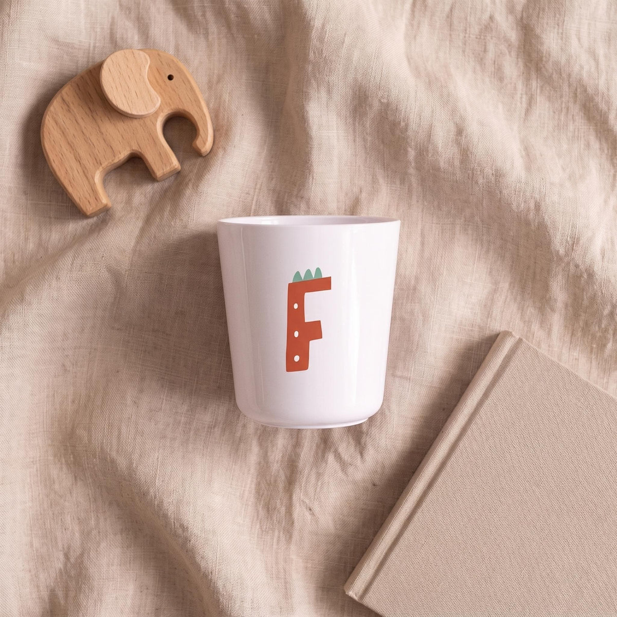 A white plastic cup featuring a red letter F decorated like a dinosaur, placed on a neutral wrinkled fabric surface next to a wooden toy elephant and a closed beige book.