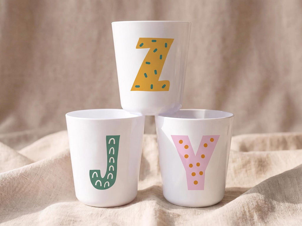 Three white plastic cups stacked in a pyramid shape on a beige fabric background, with each cup displaying a different patterned alphabet letter in yellow, green, and pink.