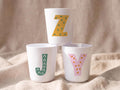 Three white plastic cups stacked in a pyramid shape on a beige fabric background, with each cup displaying a different patterned alphabet letter in yellow, green, and pink.