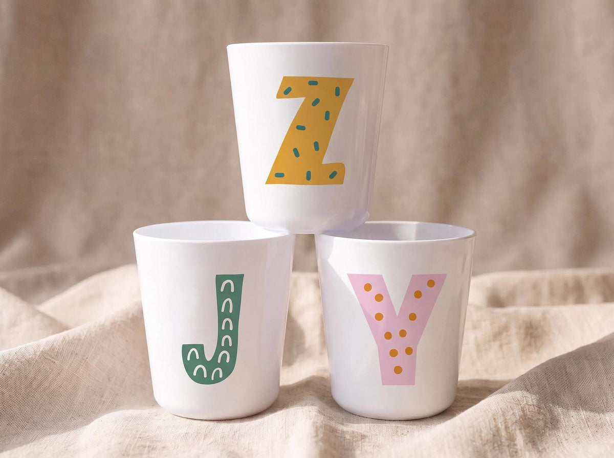 Three white plastic cups stacked in a pyramid shape on a beige fabric background, with each cup displaying a different patterned alphabet letter in yellow, green, and pink.