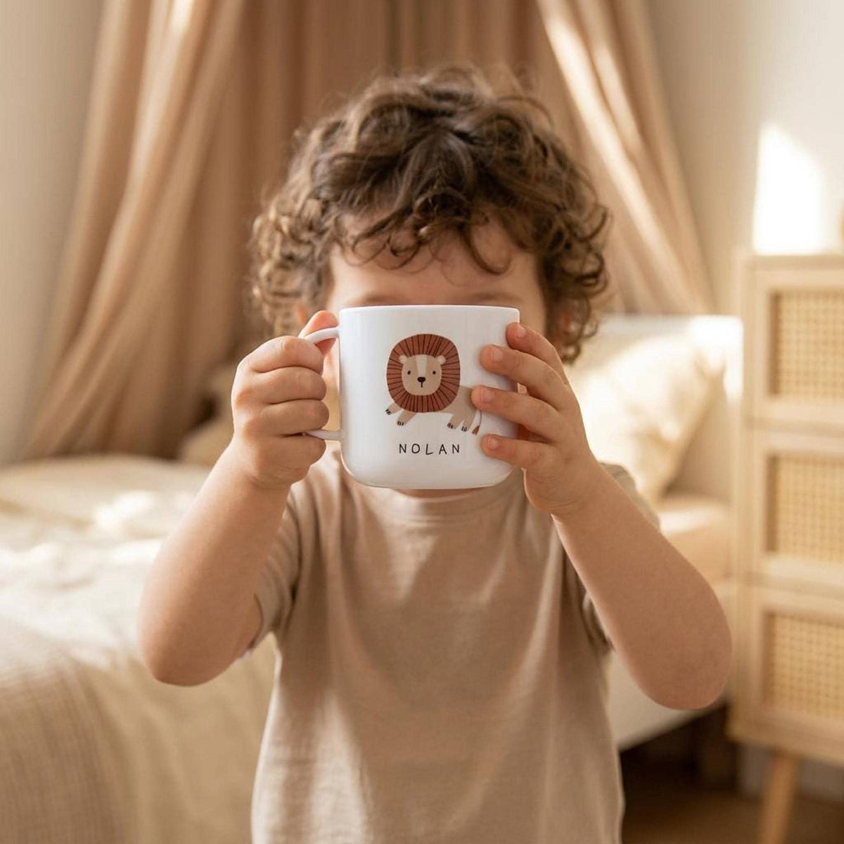 A toddler holds a white mug with a lion illustration and the name Nolan in front of their face inside a bedroom.
