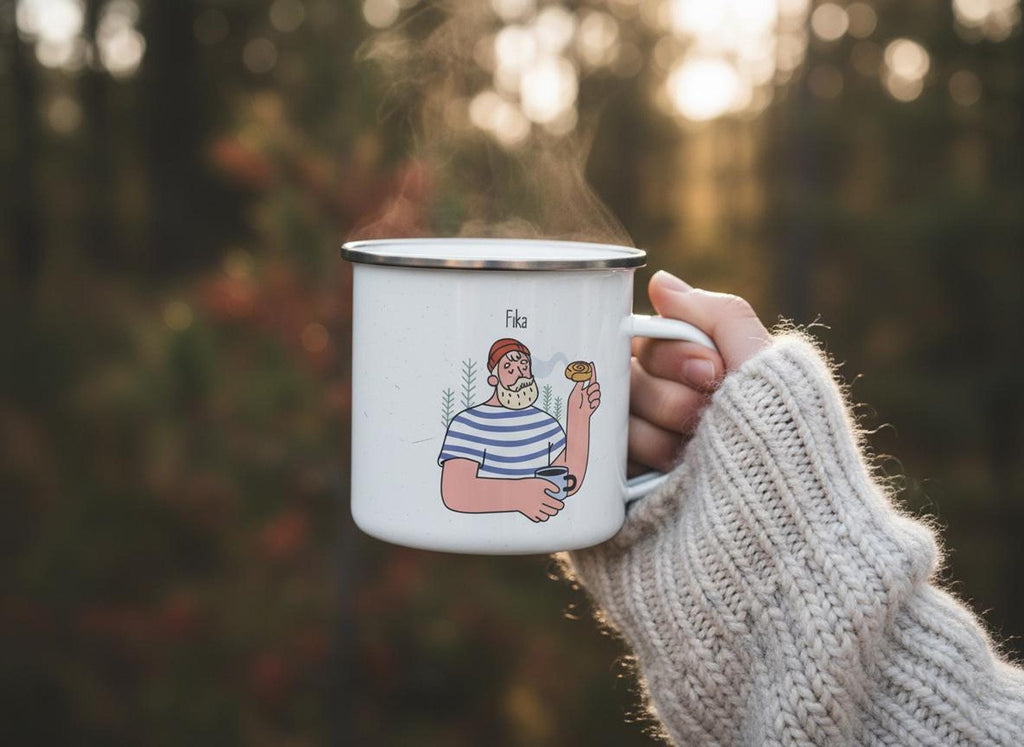 A hand wearing a cream-colored knit sweater holds a steaming white enamel mug with the man and pastry Fika illustration outdoors.