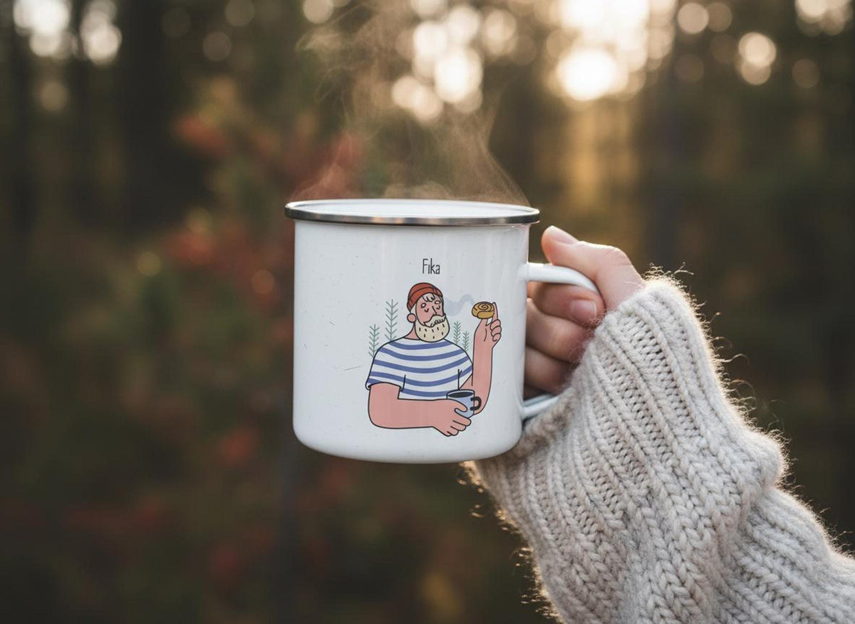 A hand wearing a cream-colored knit sweater holds a steaming white enamel mug with the man and pastry Fika illustration outdoors.