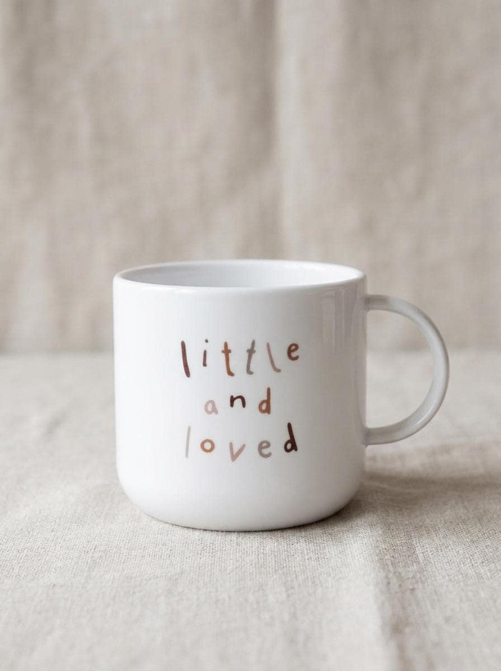 A plain white children's mug featuring the text little and loved in brown letters, resting on a textured beige fabric.