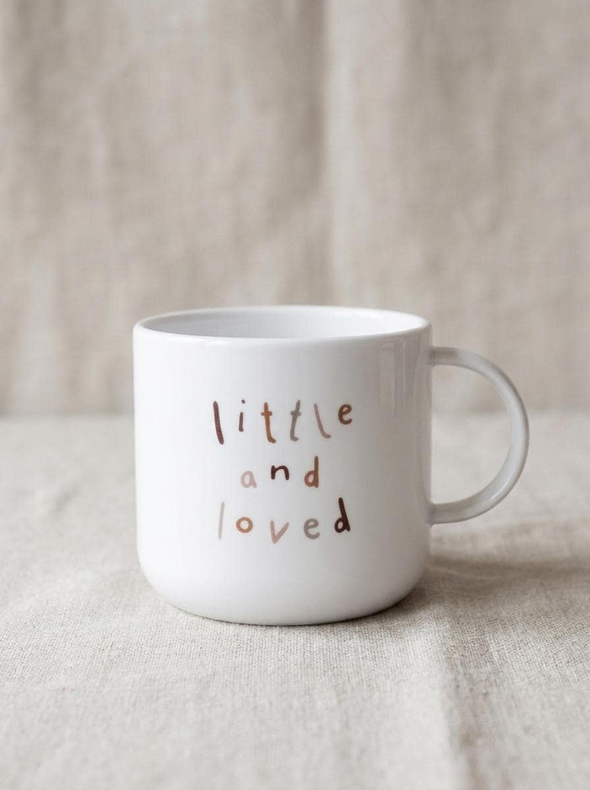 A plain white children's mug featuring the text little and loved in brown letters, resting on a textured beige fabric.