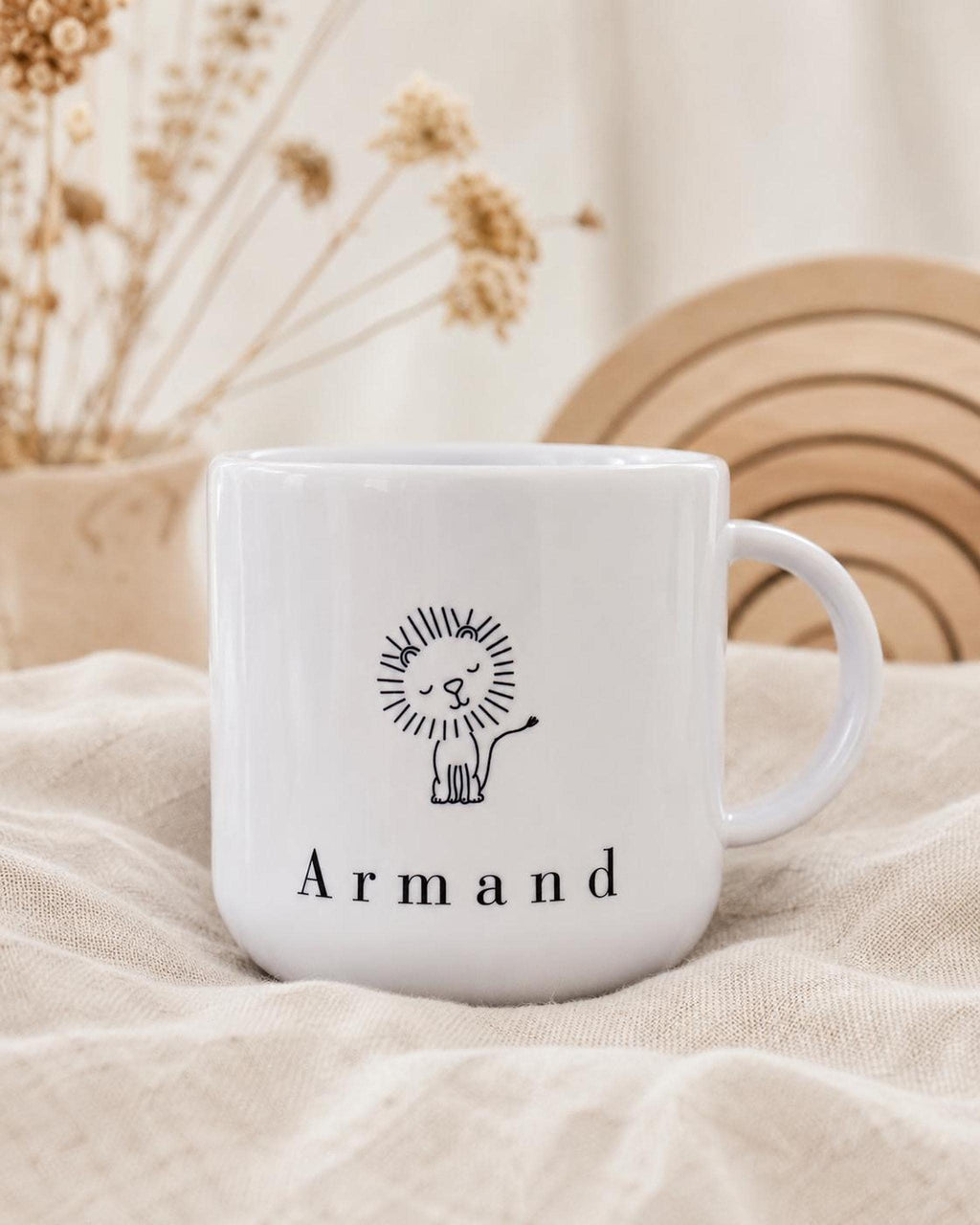 The white personalized kids mug with a lion graphic and the name Armand sits on draped fabric with dried florals in the background.