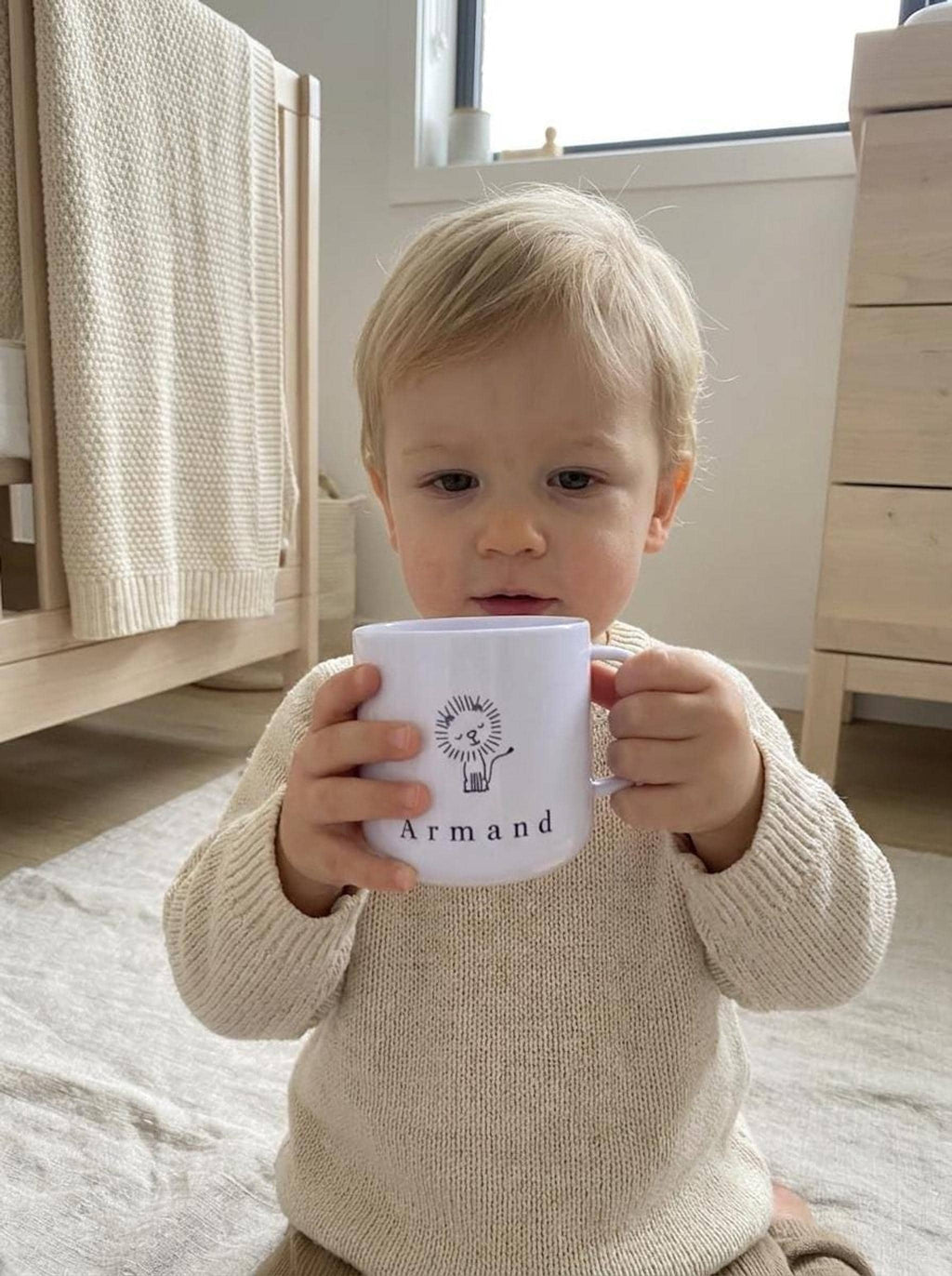A young toddler wearing a cream knit sweater holds the personalized white lion cup with both hands while sitting on a rug.