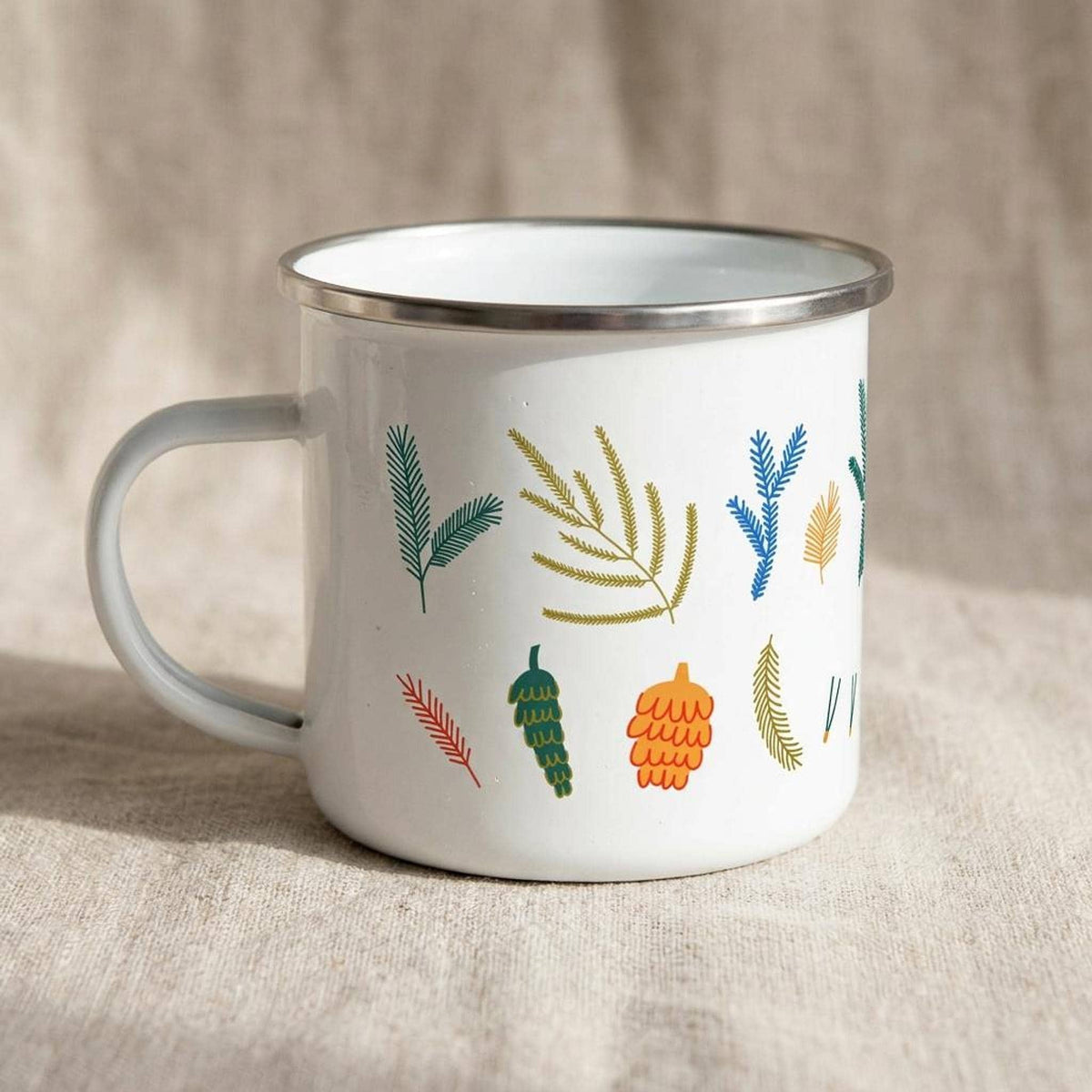 A white enamel mug with colorful botanical illustrations of pine needles and cones sits on a linen surface.
