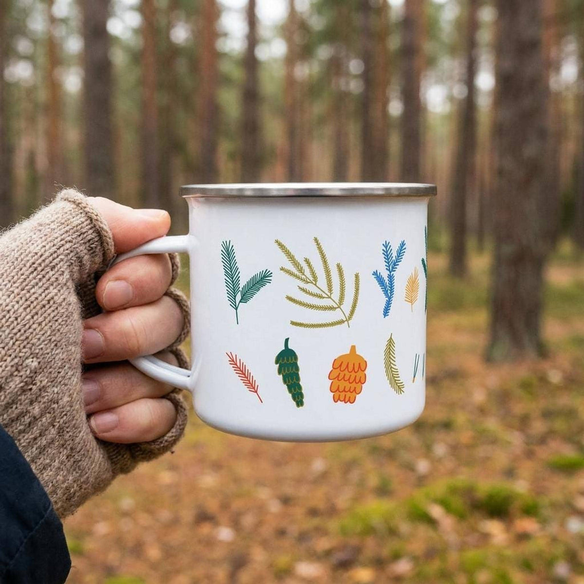 A hand wearing a knit fingerless glove holds the white botanical enamel mug in front of a forest background.