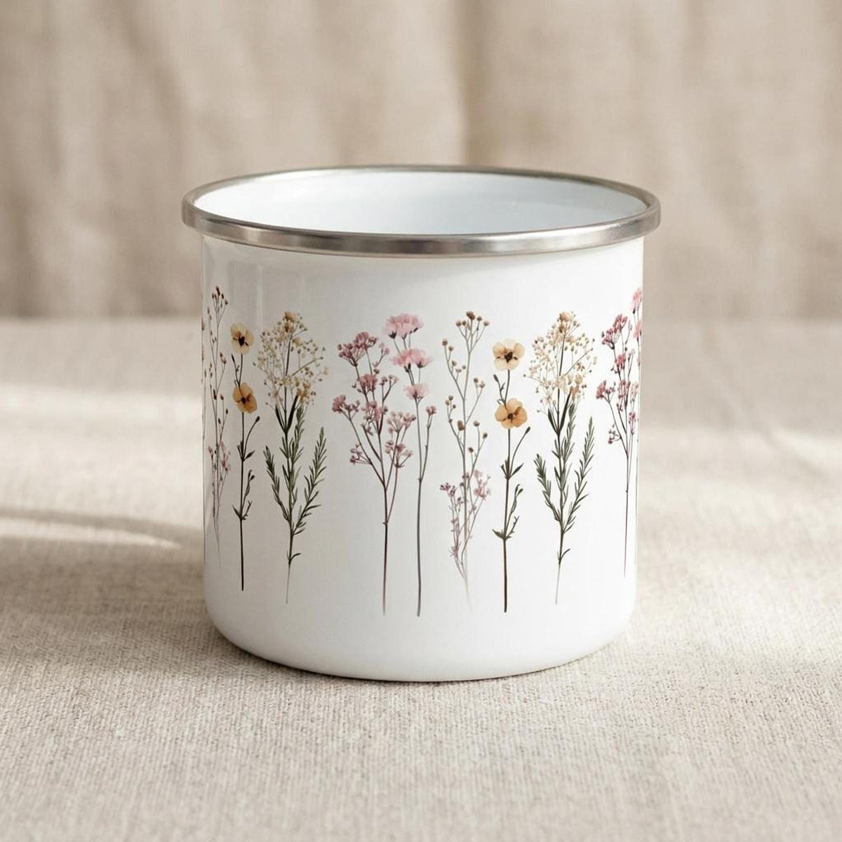 The white enamel mug is shown from another angle, displaying the repeating meadow flower graphic against the neutral fabric backdrop.